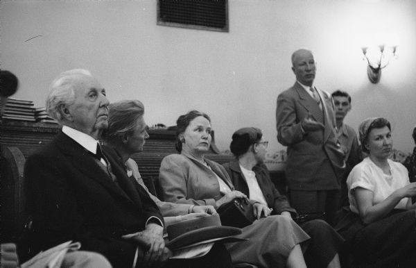 Architect Frank Lloyd Wright and others seated at the Wisconsin State Capitol during a hearing on the Frank Lloyd Wright Foundation. An unidentified man is speaking. The hearing was held in the Education and Transportation Offices, room 314 NW of the Capitol. Wright testified before the Assembly Taxation Committee in favor of a bill granting tax-exempt status to Taliesin as a non-profit education institution.