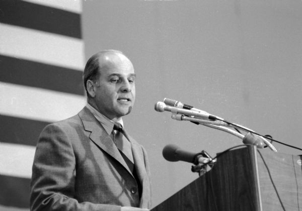 Gaylord Nelson speaking at a podium on Earth Day with a flag partially visible in the background.