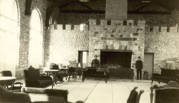 Interior view of the Thordarson library featuring a massive fireplace with two men posing on either side of it. The building is of stone, with arched windows.