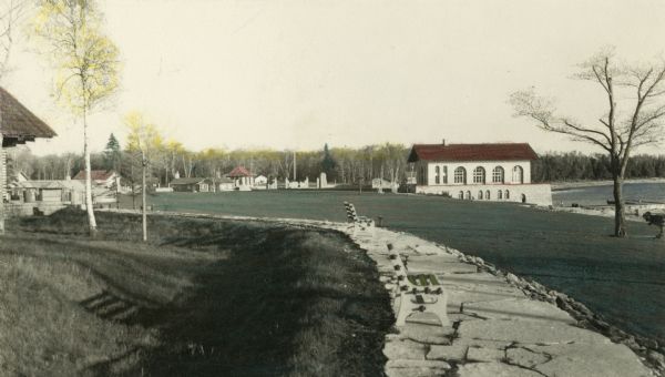 Hand-colored view of the Thordarson estate including the boathouse and a stone-paved path with benches.