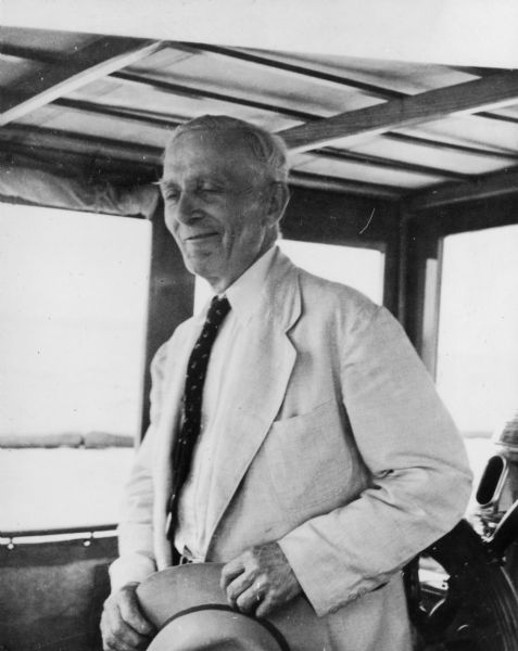 Candid portrait of Chester Thordarson wearing a light-colored suit, holding a hat. He appears to be on a boat, with windows and a steering wheel behind him.