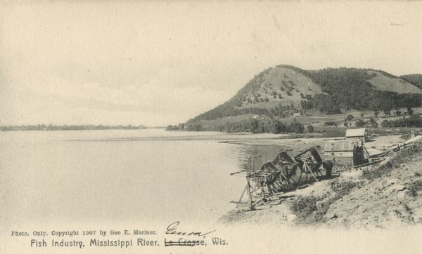 Fishing implements on the shoreline of the Mississippi River, and a bluff in the background. Caption reads: "Fish Industry, Mississippi River, La Crosse, Wis." ["La Crosse" has been crossed out, and Genoa has been written in.]