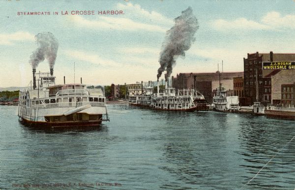 Several steamboats in the harbor, with J.J. Hogan Wholesale Grocery store and other buildings along the shoreline. Caption reads: "Steamboats in La Crosse Harbor."