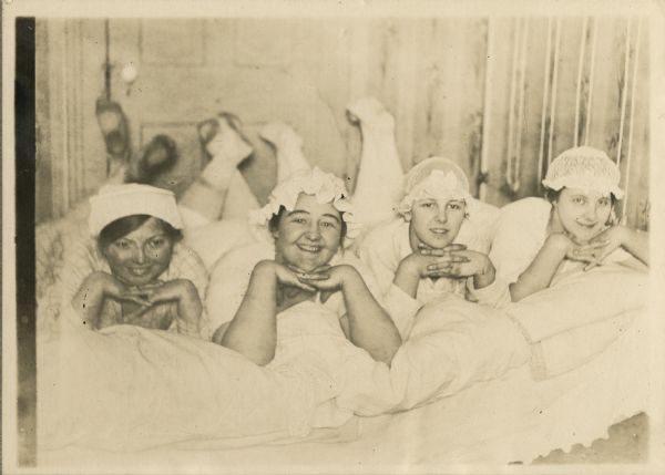 Four women, dressed in white nightgowns and nightcaps laying on a bed.