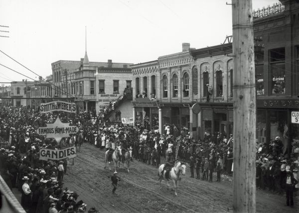 Crowds of individuals assembled on the sides of the street to watch parading soldiers, and Native Americans riding horses. Looking east on Main Street from between 2nd and 3rd Streets.