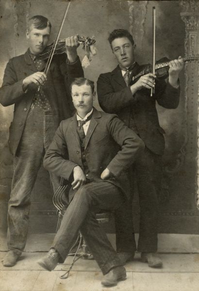 Three men, two of them standing with string instruments and the other seated.