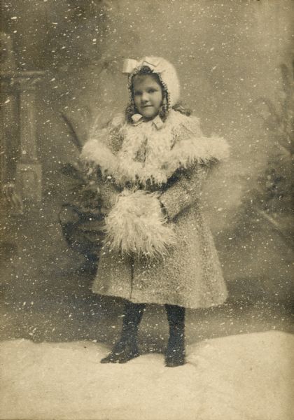 Portrait of a young girl, clothed in a winter coat, bonnet, and muff, with fake snow on the ground and falling all around her.
