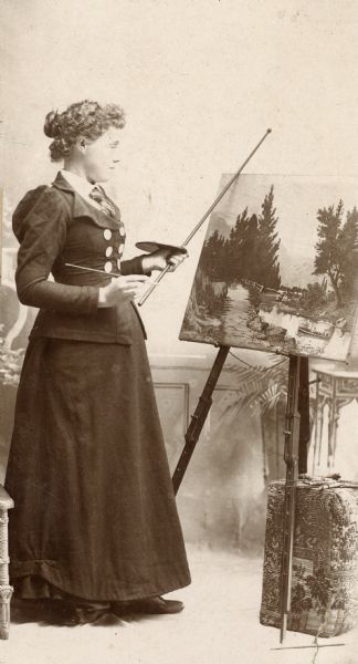 A woman holds a paintbrush, palette, and painter's stick while observing her painting on an easel in front of her.