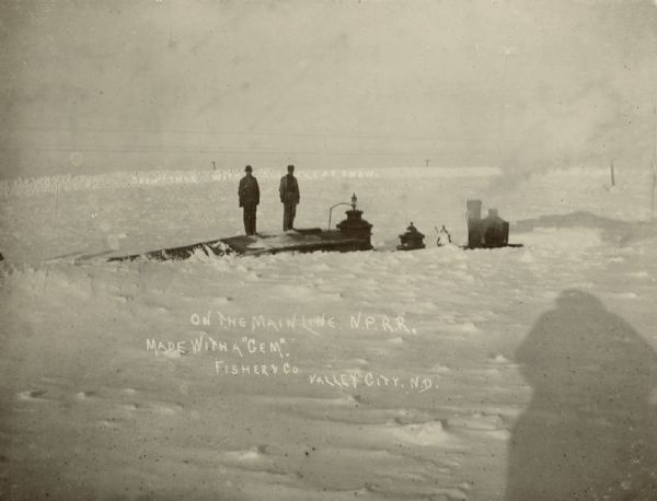 Two men standing on top of a snow-covered railroad car. The top of the steam locomotive is the only area visible of the train. Photograph reads, "On the Main Line, N.P.R.R. [North Pacific Rail Road]." "Made with a Gem," the Gem possibly being the type of camera used to take the photograph.