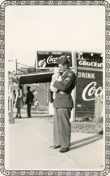 A World War II soldier holds an infant in front of a grocery store.