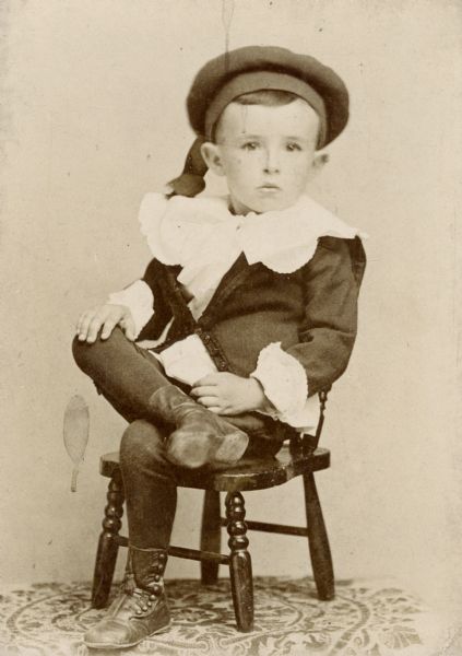 A young boy poses in a costume from the late nineteenth century, while sitting on a small chair.