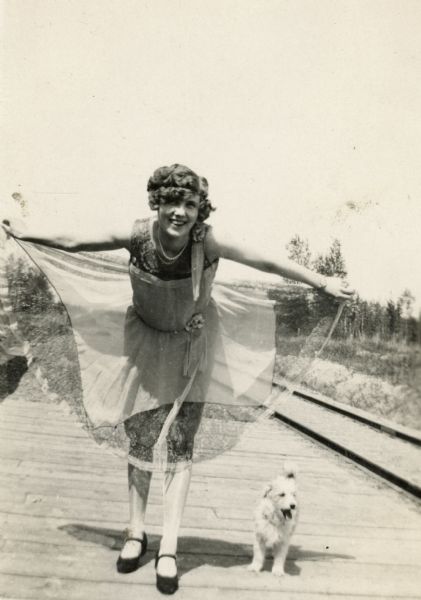 A woman curtsies towards the camera with a little dog to her left.