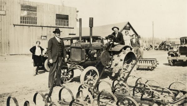 Group in a John Deere Farm Equipment yard. Three women are in the background and two men in the foreground, one on top of the tractor.