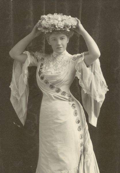 Studio portrait of Ella Wheeler Wilcox wearing an elaborate light-colored dress, with her hands on her hat.