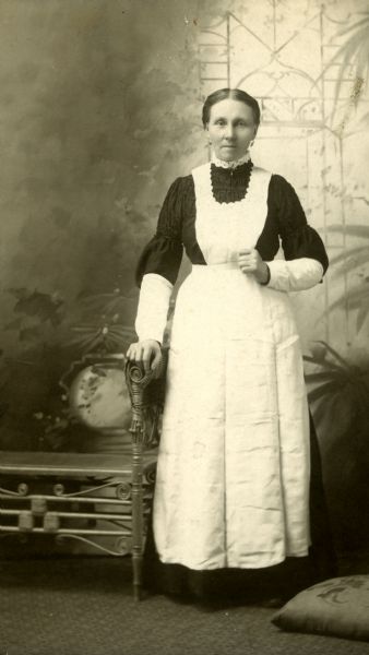 Studio portrait of Anna Berg wearing a black dress and a long, white apron.