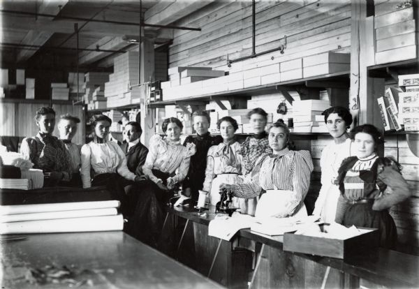 Women workers inside a textile factory, possibly Beherhausshict Factory. Clothing boxes and spools of thread are visible.
