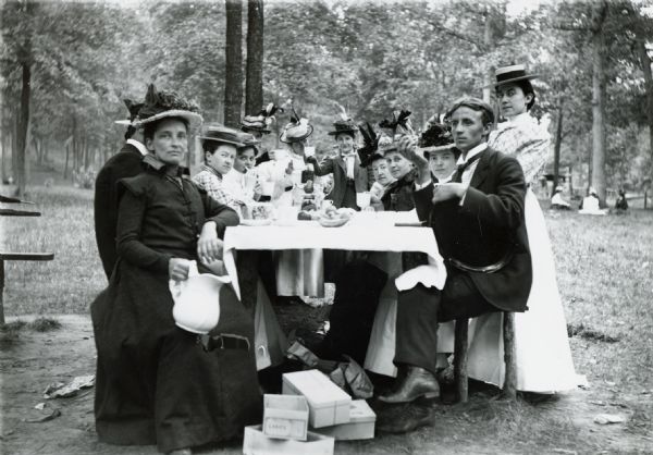 Men and women drinking tea around a picnic table at a park. The women are wearing fancy hats.