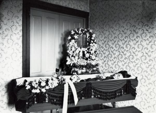 Open coffin, decorated with flowers, and a wreath that reads, "Our Mother" in a funeral salon.