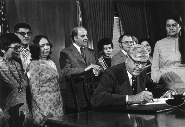 Secretary of the Interior Rogers Morton signing the documents that restored tribal status to the Menominee Indians. Watching the ceremony are Senator Gaylord Nelson (directly behind Morton), and Ada Deer, head of the interim Menominee Restoration Committee (standing at the right).