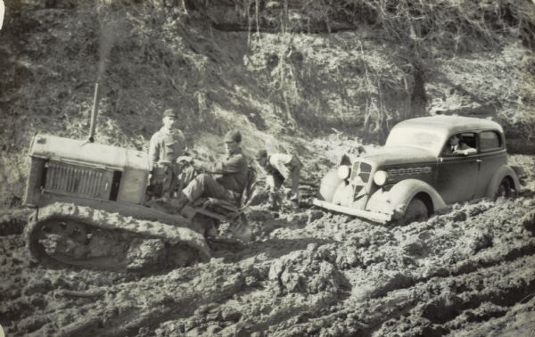 Men with a tractor attempt to pull out a car stuck in the mud on an early road.