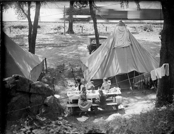 Elevated view of the Herman Taylor family camping at Devil's Lake. There is a moving train in the background on railroad tracks along the shoreline of the lake.