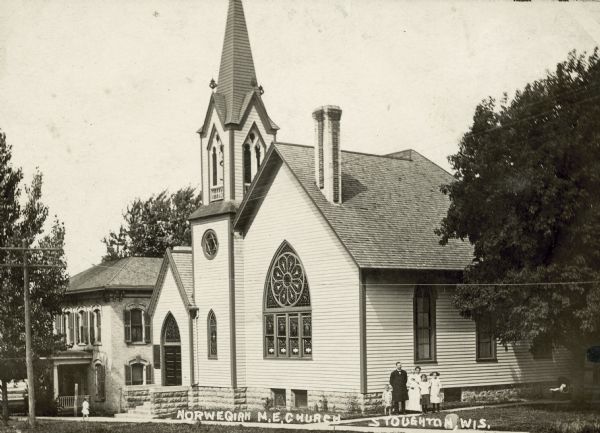 Exterior view of the Norwegian Methodist Episcopal Chruch. There are five people, probably a family, posing in front of the building. Caption reads: "Norwegian M.E. Church, Stoughton, Wis."