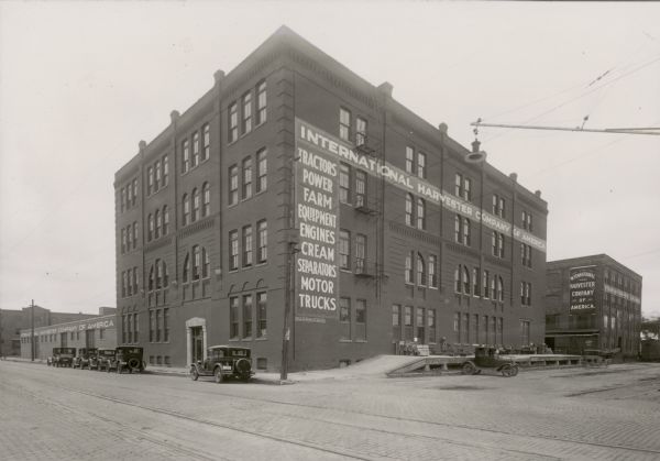Street scene with several buildings painted with the words "International Harvester Company of America". The building in the foreground has the words, "Tractors, Power Farm Equipment, Engines, Cream Separators, Motor Trucks" painted down the side of it, above a "General Outdoor Adv Co" plaque. Automobiles are parked at the front and side of the building. The loading dock of the building in the foreground has packing crates and large wheels on it. A ghost-like image of a moving car or wagon appears at the rear of the dock. In the left background is a building with the letters "NNSYLVA" visible. 
