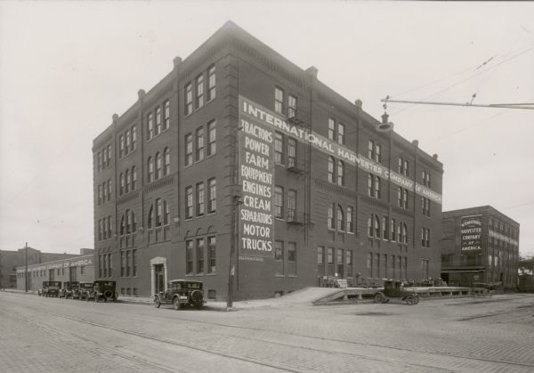 Street scene with several buildings painted with the words "International Harvester Company of America". The building in the foreground has the words, "Tractors, Power Farm Equipment, Engines, Cream Separators, Motor Trucks" painted down the side of it, above a "General Outdoor Adv Co" plaque. Automobiles are parked at the front and side of the building. The loading dock of the building in the foreground has packing crates and large wheels on it. A ghost-like image of a moving car or wagon appears at the rear of the dock. In the left background is a building with the letters "NNSYLVA" visible.
