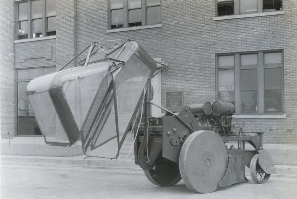 Engineering photograph of an experimental single-row, two drum, self-propelled cotton picker parks. The photograph was taken outside the office of International Harvester's Tractor Works and displays the right side of the machine with the cotton container elevated half-way to show the blower and rear cleaner. The door is released.