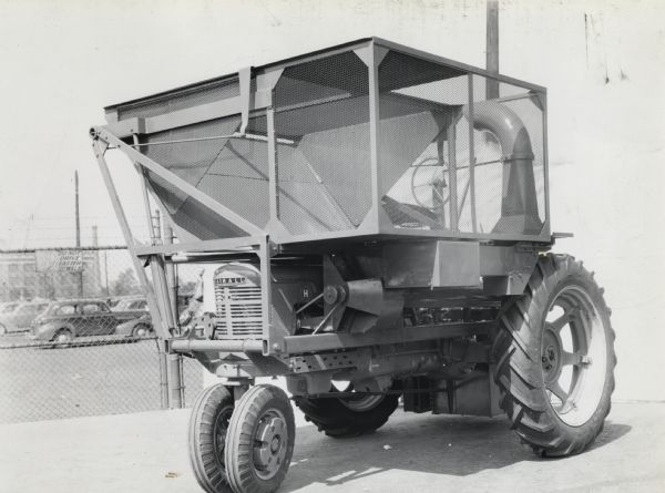 Engineering photograph of an experimental one row, two drum cotton harvester [picker] attachment mounted on a Farmall "H" tractor. The tractor is parked in front of automobiles in a fenced-in parking lot.