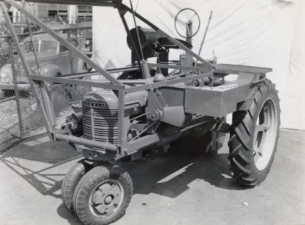 Engineering photograph of an experimental one row, two drum cotton harvester [picker] attachment on a Farmall "H" tractor, parked near a parking lot.