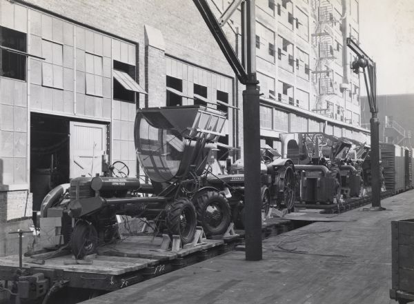 A shipment of tractor-mounted cotton pickers loaded on railroad flat cars near a factory(?) building.