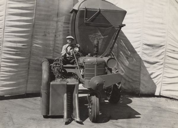 Engineering photograph of a man behind the wheel of a "1944 season" cotton picker mounted on a Farmall B tractor. There is a large canvas behind the cotton picker, perhaps for a backdrop.