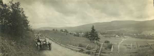 Panoramic view of a group of people in cars parked on the side of a road near a weather-worn fence. Hills and a valley are seen in the background. A town can be seen down in the valley.