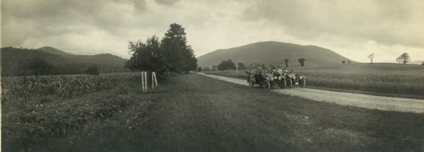 Panoramic view of a group of men and women posing with two cars side by side on a road. Hills are seen in the background.
