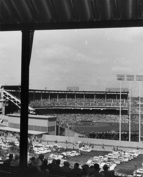 View of Milwaukee County baseball stadium showing parking lot in foreground and including view of stadium seating and field with players.
