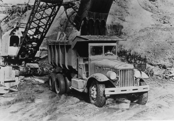 An artistically rendered photograph of a A-8 truck, with a slight variation on the cab. The A-8 is being used as a dump truck at an excavation site; a crane dumps dirt into the open-box bed.