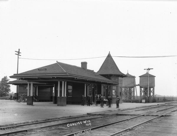 Exterior view of the Corliss train depot with railroad tracks in the foreground. A small group of men pose in front of the building.
