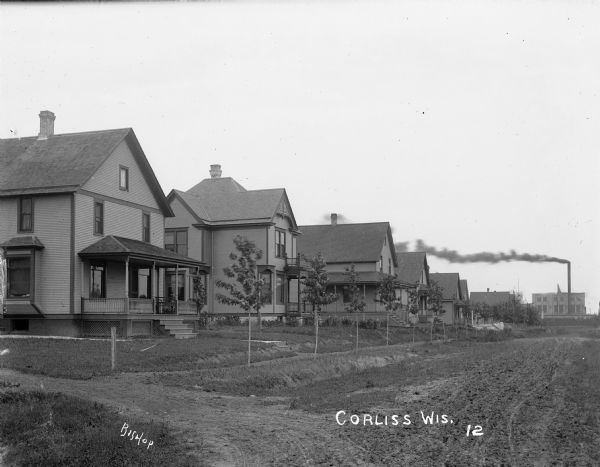View down a dirt road with houses lining the street. A factory is visible in the distance.