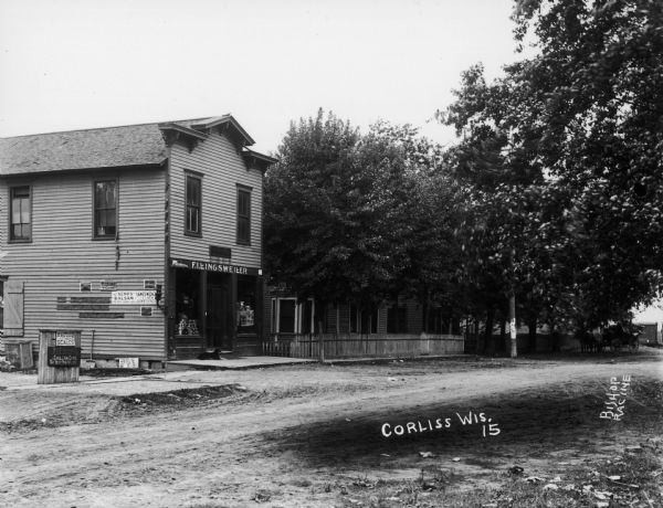 A building on a main street in Corliss, which may be a general store. There are many advertising signs on and near the building. A dog lies on the front steps.