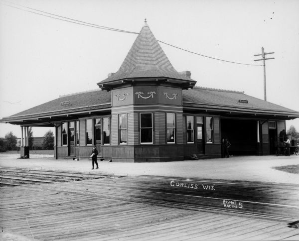 Train depot with a boy standing between the building and the railroad tracks. There are two doors marked "Men" and "Women". Four men can be seen on the right side of the depot.