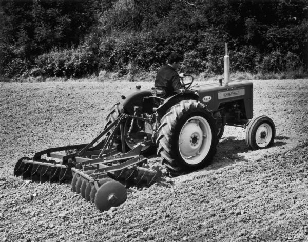 A man sits at the wheel of a British B-414 tractor in a field. The tractor has a disk harrow attached.