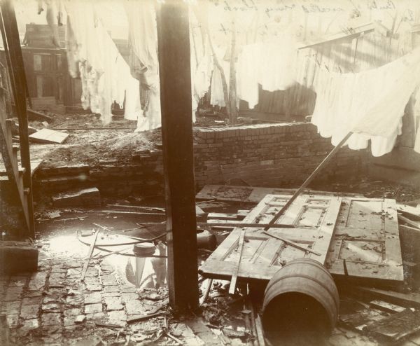 A slum courtyard on 40 Albemarle Street, with laundry hanging to dry. There are broken doors and a barrel on the ground.