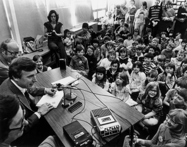 Governor Martin Schreiber is seated at a desk surrounded by children. The photo was of the signing of an education bill.  Senator Fred Risser is to Schrieber's left at the table. The woman to the Governor's right was the then Superintendent of Public Instruction.
