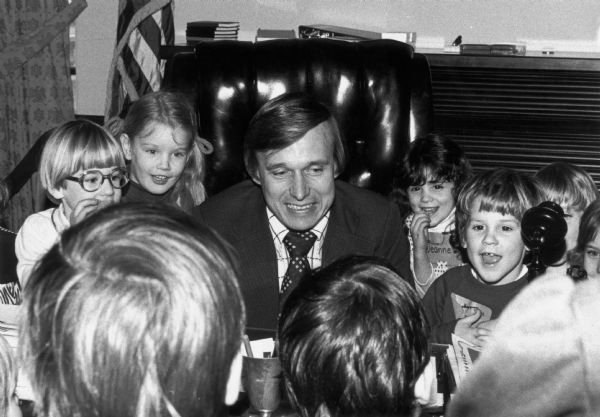 Governor Martin Schreiber is seated at a desk surrounded by children.