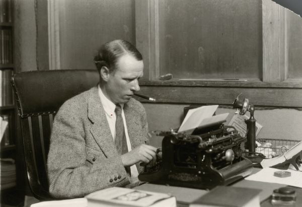 Sinclair Lewis seated at a typewriter with a cigarette in a holder in his mouth.