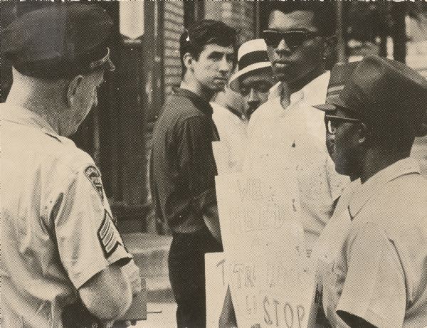 Tom Hayden, in the center wearing the dark shirt, looking at a police officer talking to four African-American men. Newark ERAP project. From the SDS collection.