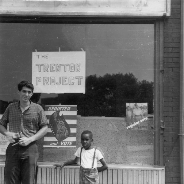 Tom Hayden standing in front of building with a small African-American child. Poster on window for "The Trenton  Project" and two posters advocating that people register to vote. From the SDS collection.