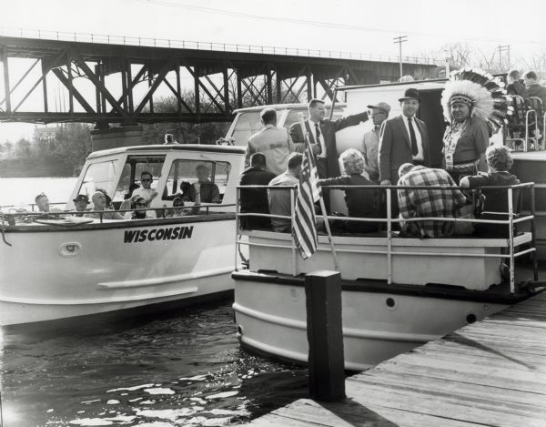 Anthony Wise in a necktie and hat standing next to a Native American wearing a feather headress, on a boat adjacent to a dock.
