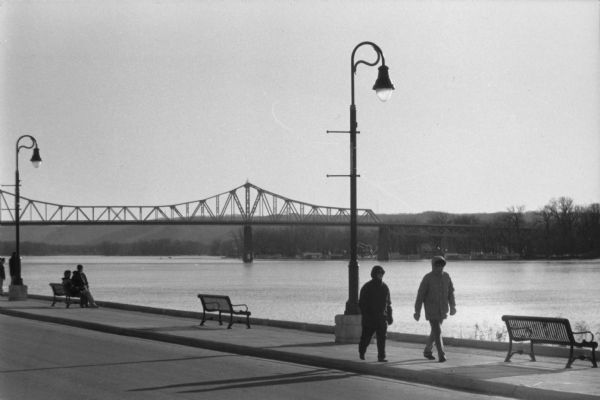 A couple walking along the riverfront in winter, with a bridge across the Mississippi in the background.